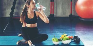 Woman in workout clothes drinking water next bowls of fruit and vegetables